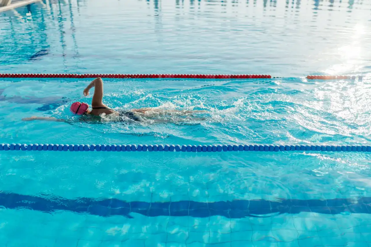 A swimmer in a pink cap glides through the blue water, performing a recovery swim workout. Sunlight glints off the surface.