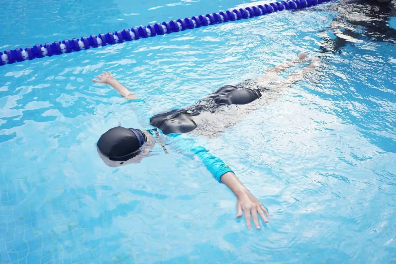 A person wearing a black swim cap and a black swimsuit is floating in a bright blue swimming pool. One arm, in a blue sleeve, is extended.