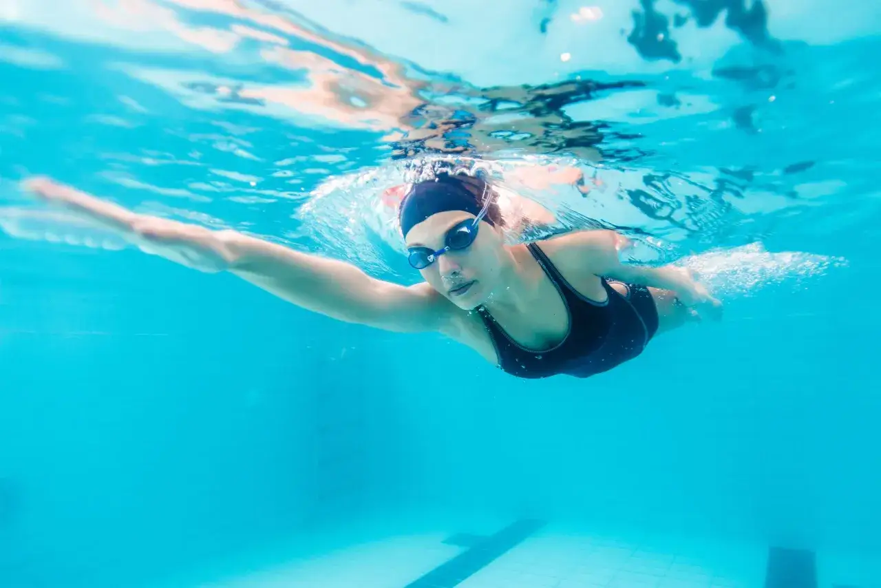 Underwater shot of a woman swimming laps. She wears a black cap and goggles. This image can represent recovery, asking 
