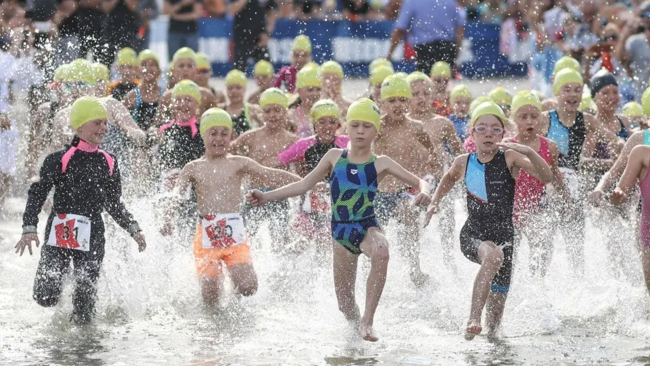 Children in yellow caps splash through the water at the start of a youth triathlon.