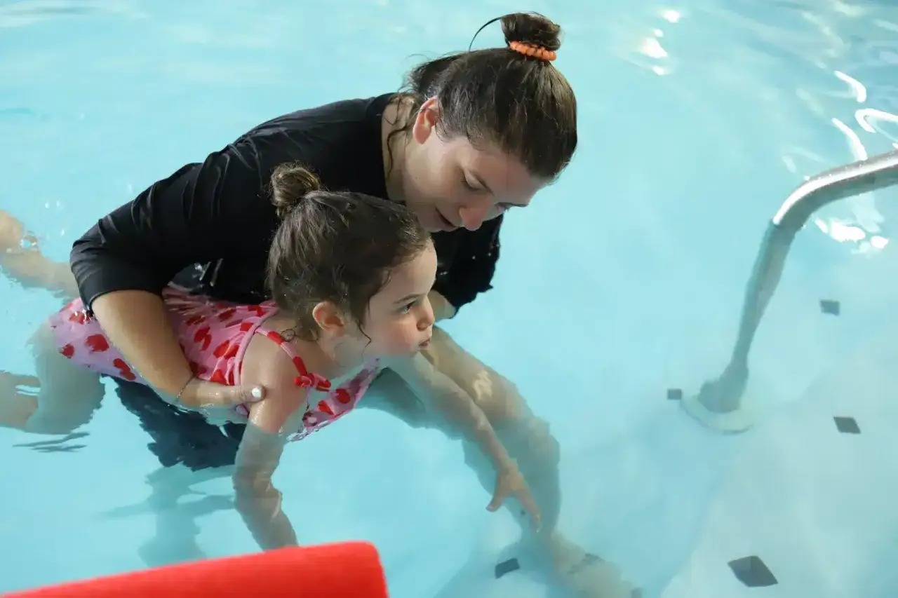 A woman gently holds a toddler in a pool, demonstrating how to teach a toddler to hold breath underwater.