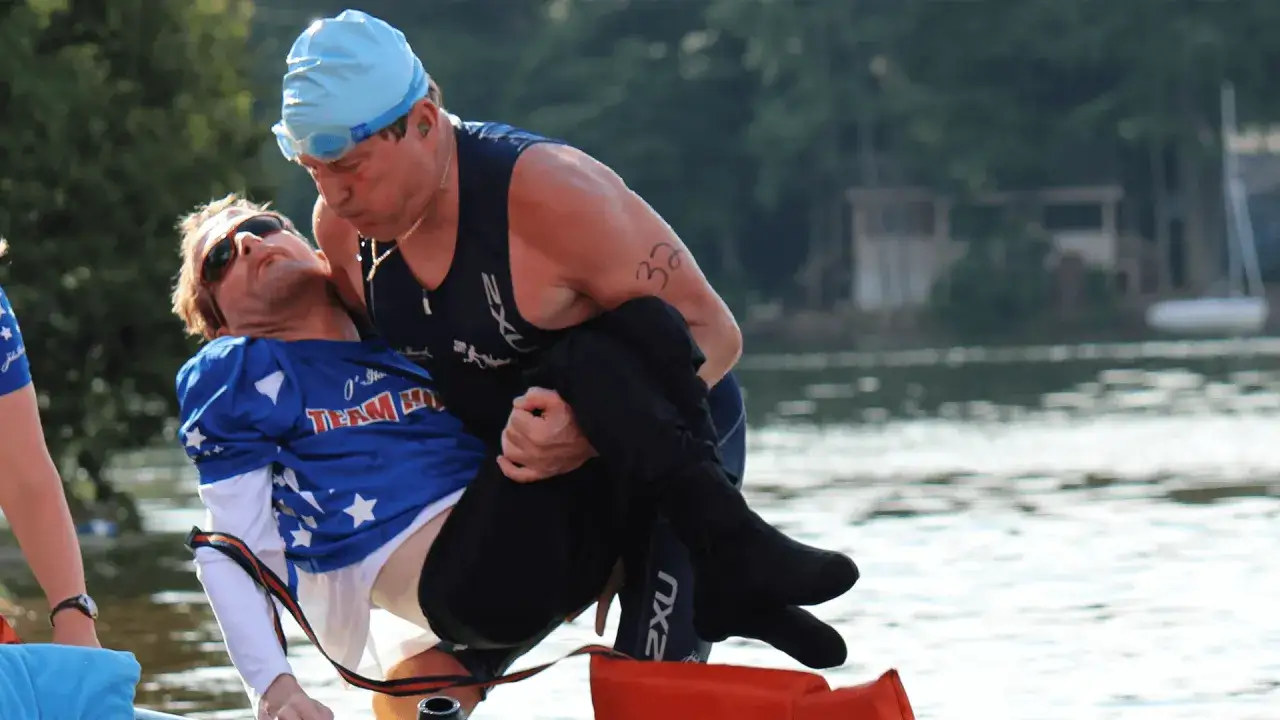 A man in a blue cap and goggles carries another man in a blue shirt with stars, representing the spirit of Dick Hoyt and Ironman.