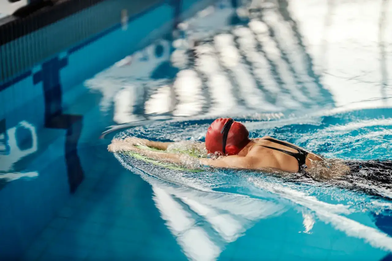 A swimmer in a red cap and black suit uses a kickboard, their body submerged in the blue pool. The resistance of the water makes the pool a great workout, explaining why does the pool make you tired.
