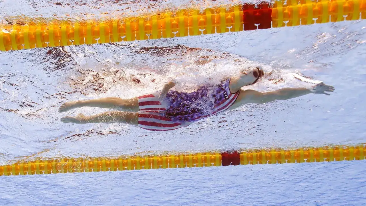Underwater shot of Katie Ledecky training, her powerful freestyle stroke propelling her through the water. Her red, white, and blue suit is visible.
