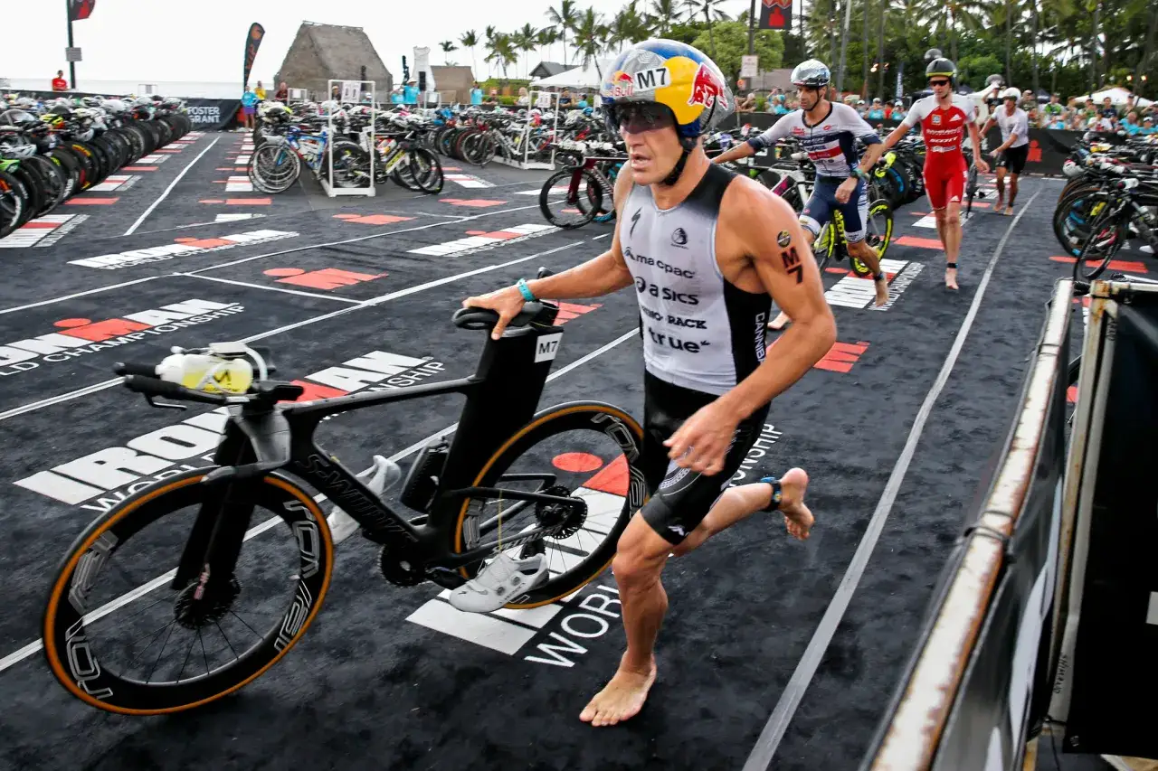 Athlete runs with his bike, ready for the full Ironman distance. Rows of bikes and palm trees in the background.