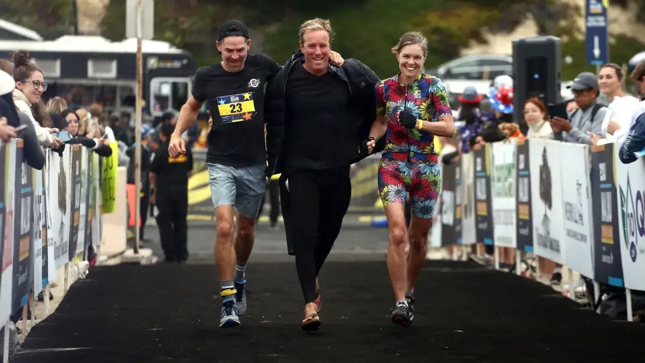 Three friends celebrate crossing the finish line of an Ironman relay. The woman in the colorful floral suit beams, while the two men, one in a black shirt with a race bib, share a laugh.