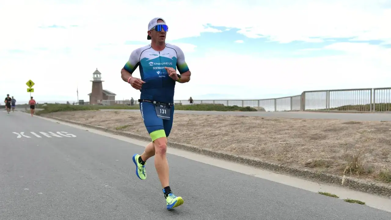A man in a triathlon running suit, bib 131, races along a coastal road with a lighthouse in the background.