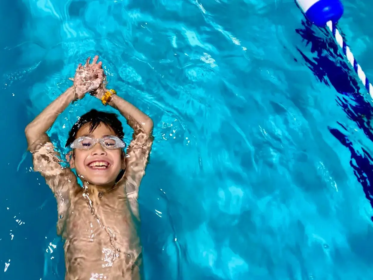 A happy boy with goggles floats on his back, his face just above the water. His arms are raised, and he's smiling widely.
