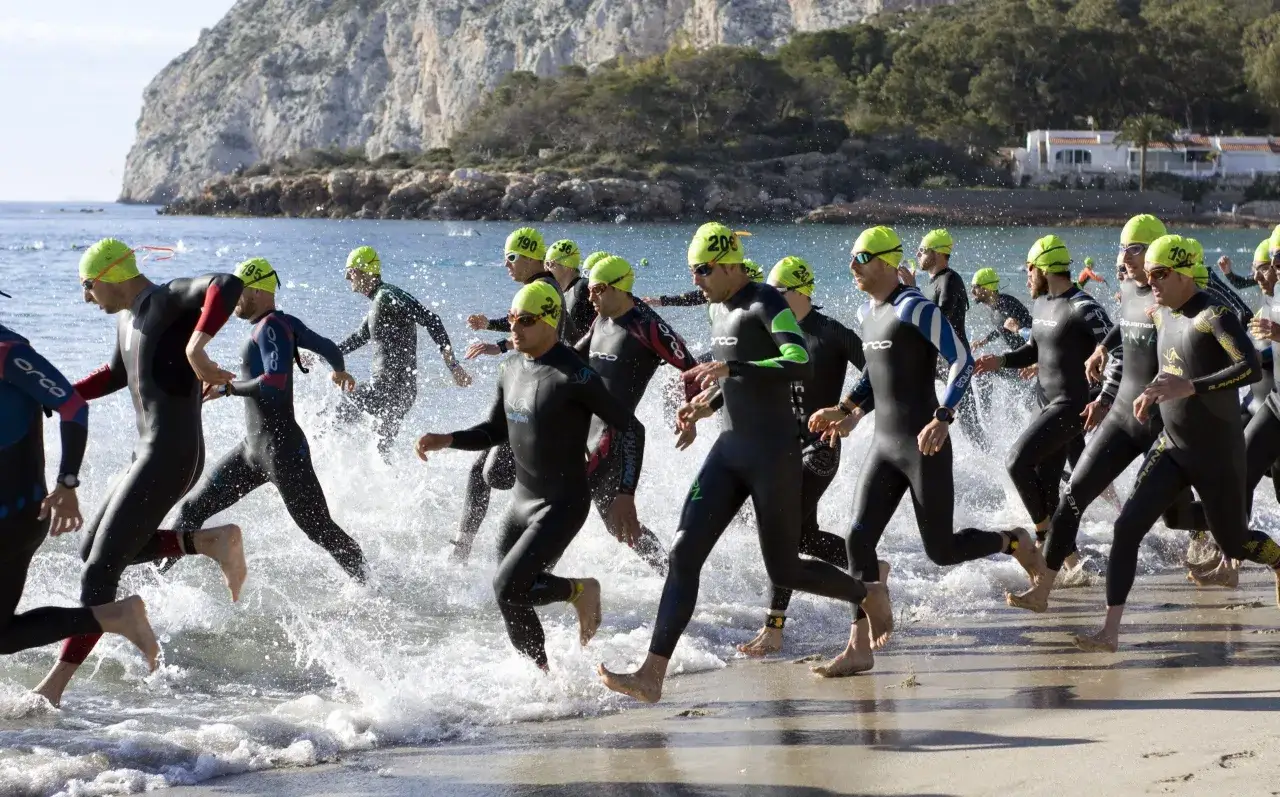 Athletes in wetsuits and bright yellow caps run into the water, starting the t100 triathlon. Waves splash around their legs as they begin the swim portion of the race.