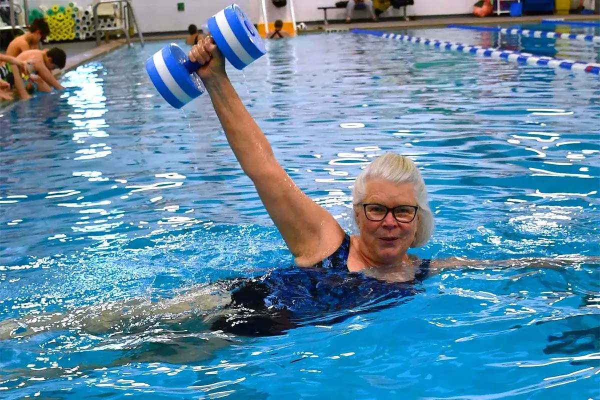 An older woman with glasses and blonde hair smiles while doing aqua zumba in a pool, lifting a blue and white water dumbbell.