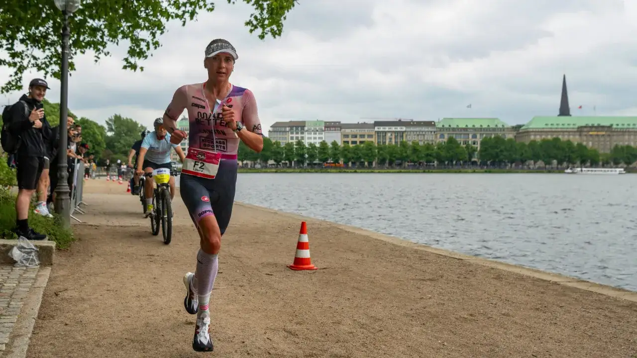 A female athlete runs along a waterfront, part of an iron man marathon length race. Buildings and trees line the distant shore.