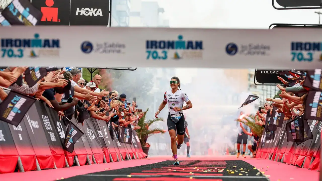 A female athlete sprints towards the finish line, crowds cheering, aiming for the fastest Ironman time.