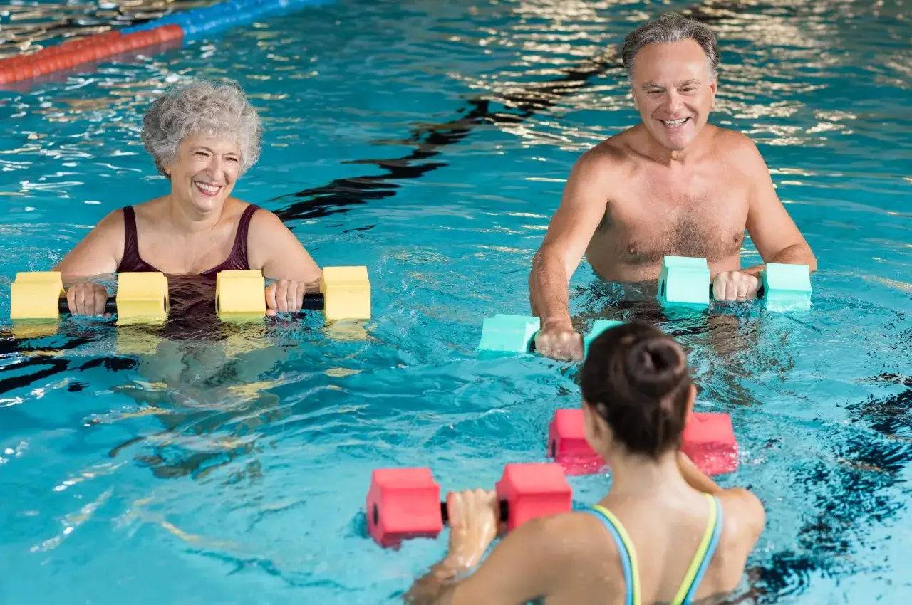 Smiling seniors and a younger woman enjoy an aqua gym class, using buoyant weights for a refreshing workout in the pool.