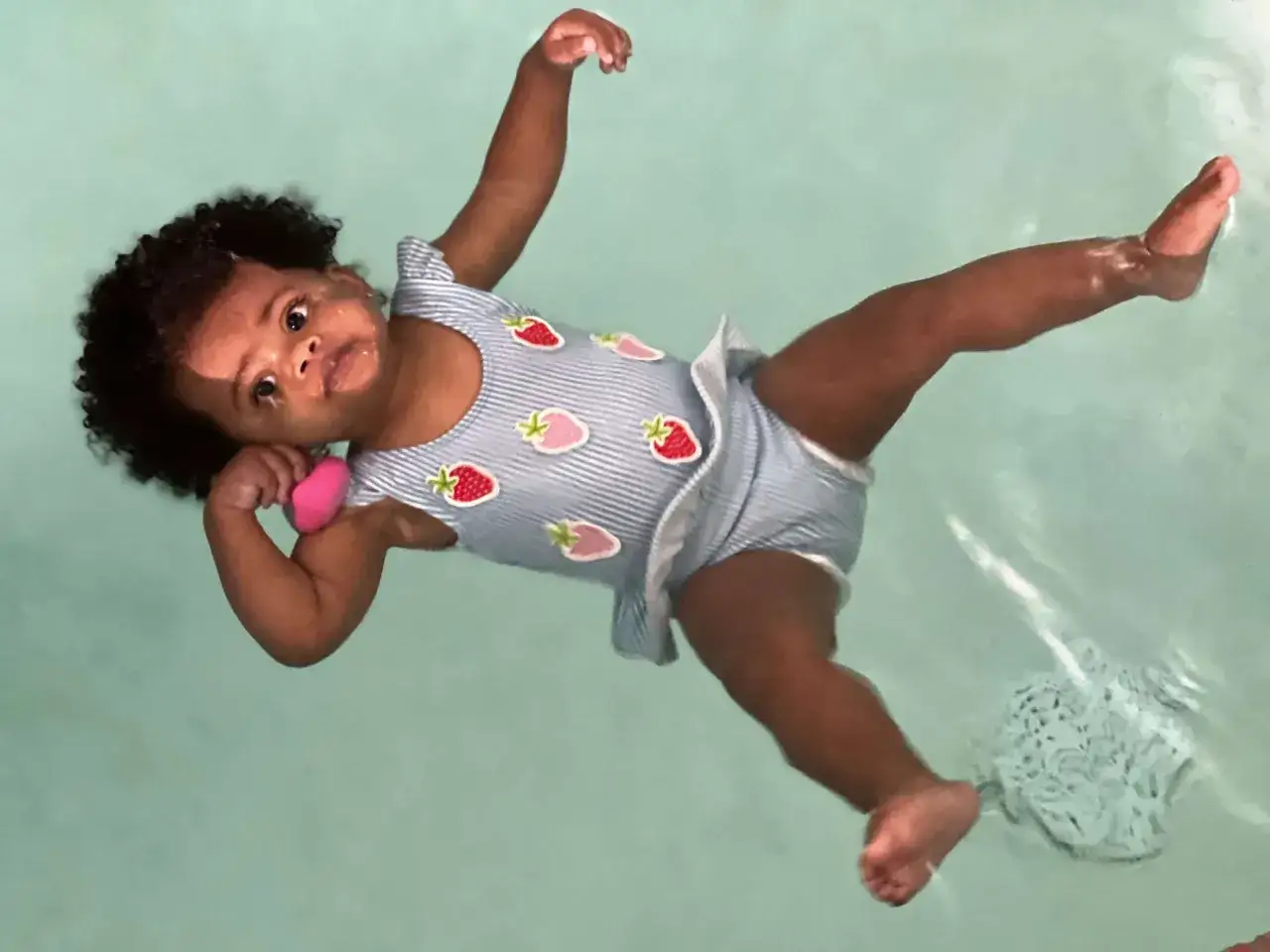 A baby in a strawberry-patterned swimsuit floats on their back in light blue water. They seem to be enjoying their ISR swim lesson, learning what ISR swim stands for: Infant Self Rescue.