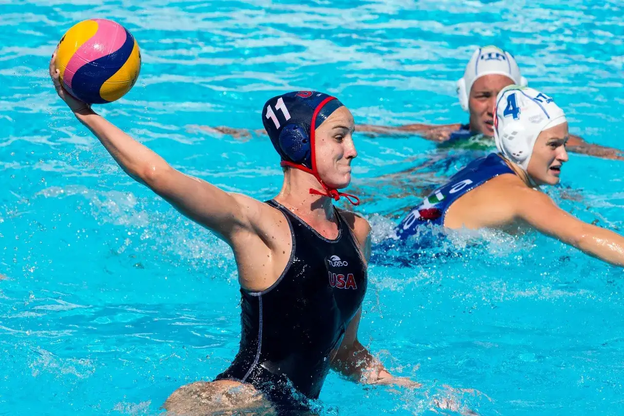 A USA water polo player with cap #11 prepares to throw the ball during intense water polo periods. Other players in white caps are in the background.
