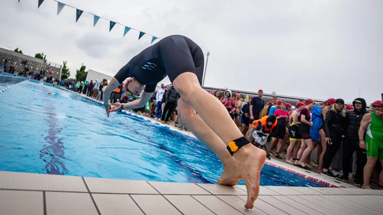 A swimmer in a black wetsuit dives into a pool, ready for advanced swim workouts. Spectators watch from the poolside.