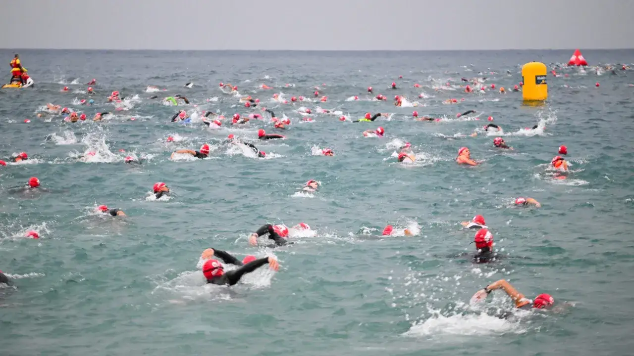 Swimmers in red caps race through choppy water during a half triathlon. A lifeguard watches from a jet ski.