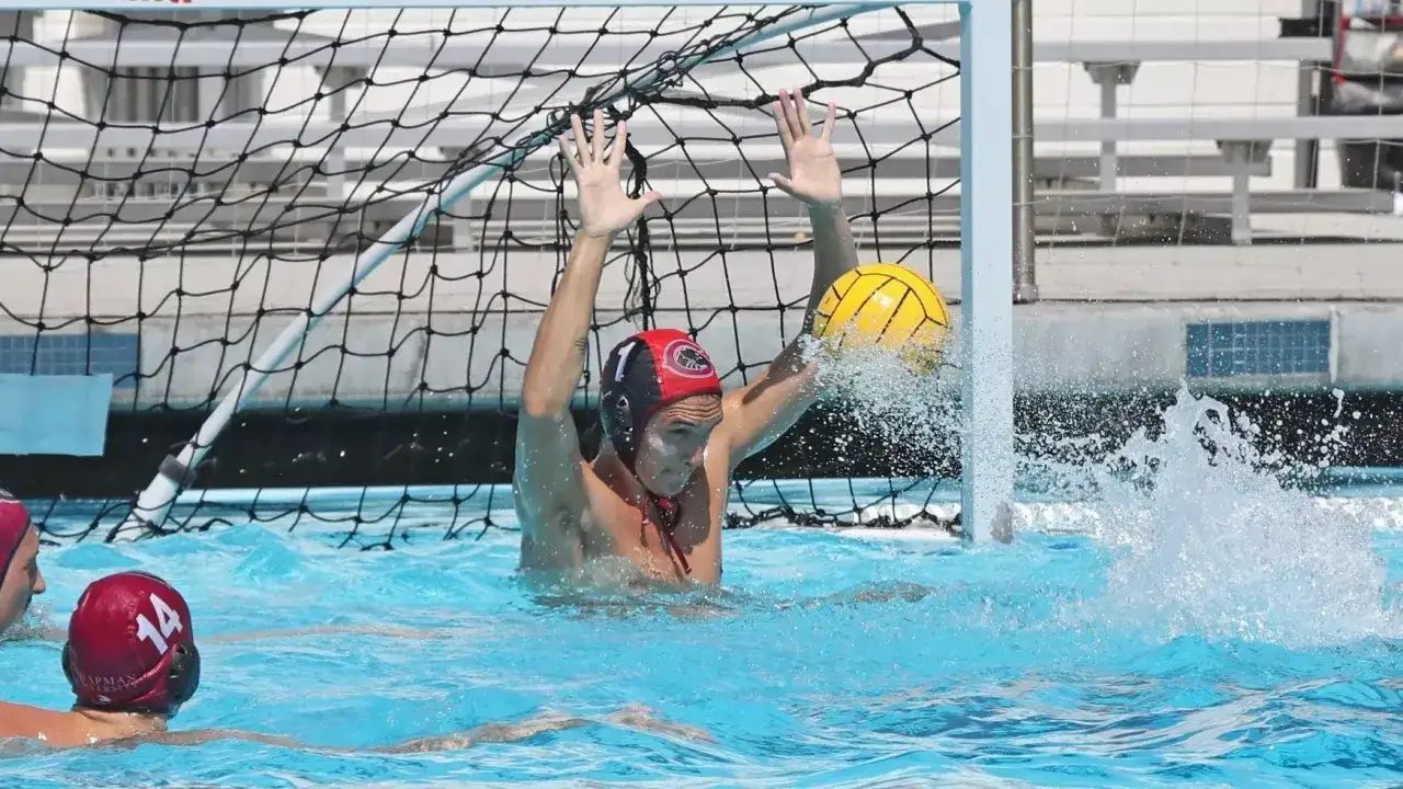 A water polo goalie with arms raised, blocking a shot. Water splashes around the goalie as the yellow ball is deflected.