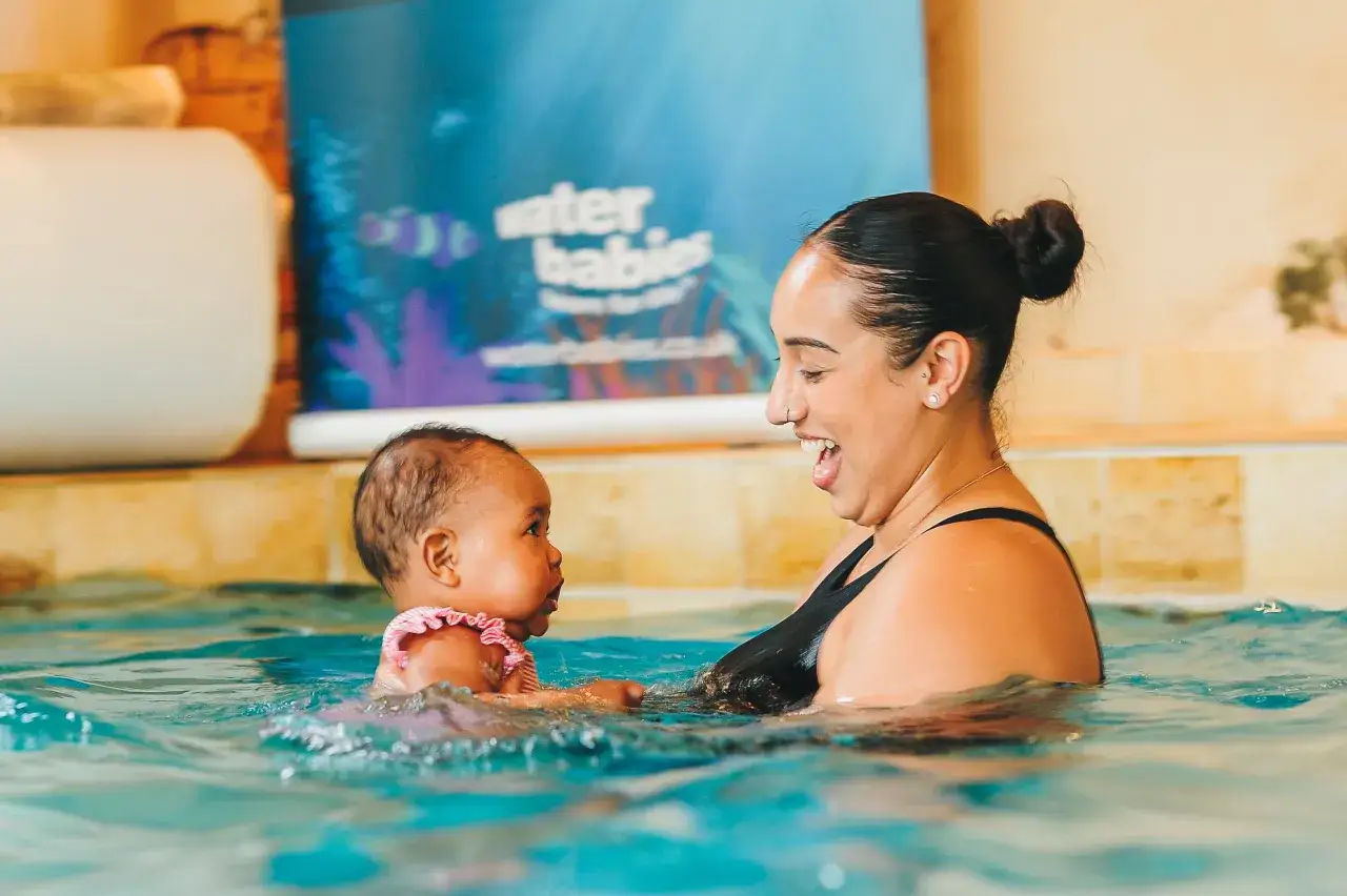 A mother laughs with her 6-month-old baby during swim lessons. The baby wears a pink swimsuit, and the mother wears a black swimsuit.