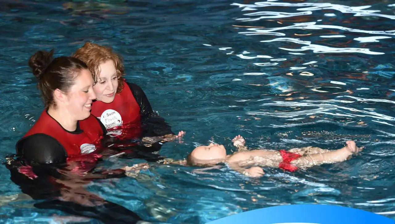 Two women in red vests teach a baby to float in a pool, demonstrating how to become a swim instructor.