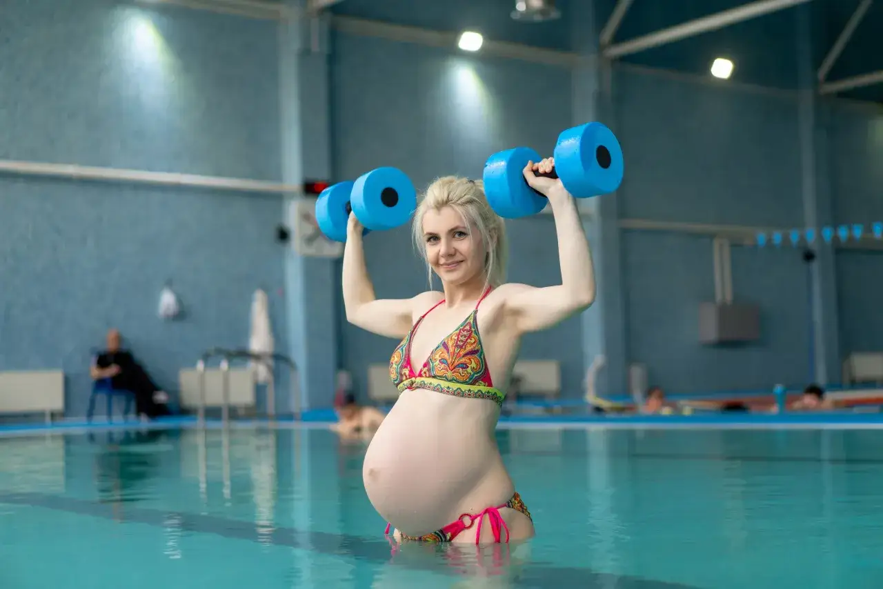 A pregnant woman exercises with blue dumbbells in a pool, showing that you can swim while pregnant.