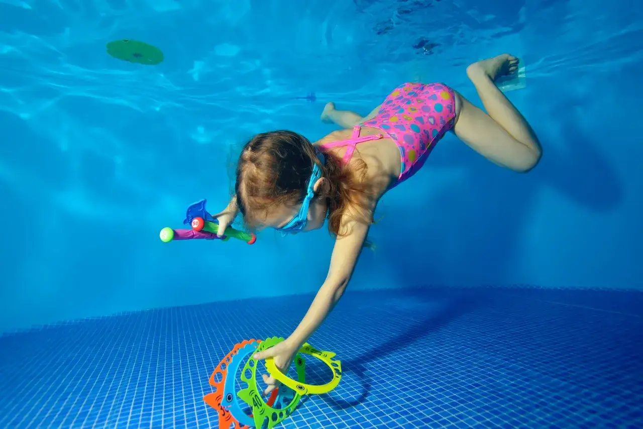 A girl in a pink swimsuit with hearts dives underwater, reaching for colorful rings, enjoying fun swim games.
