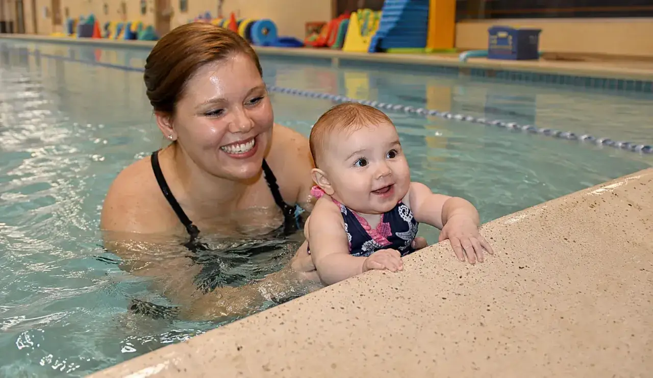 A smiling mom and baby enjoy ISR vs swim lessons in a pool. The baby, in a navy swimsuit, reaches for the edge.