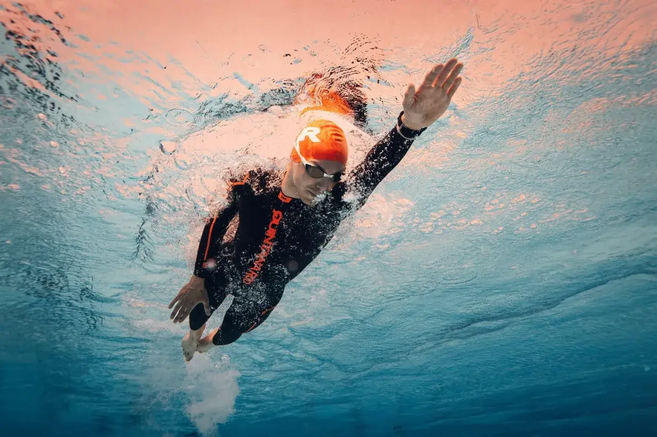 Underwater view of a triathlete performing freestyle triathlon swim strokes. The swimmer wears a black wetsuit with orange accents and an orange cap.