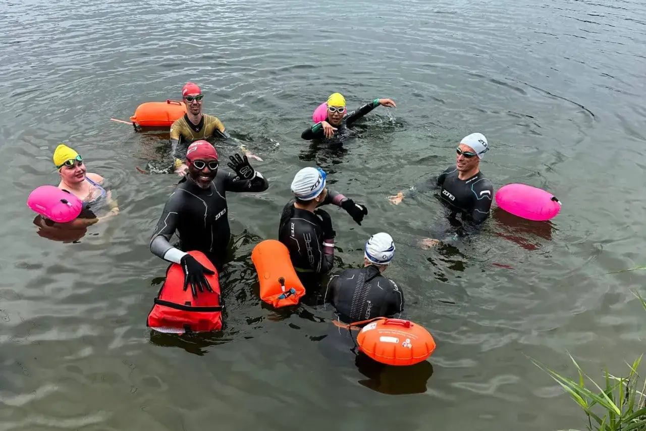 A group of swimmers in wetsuits and swim caps are enjoying their triathlon training in the water, with bright orange and pink buoys floating nearby.