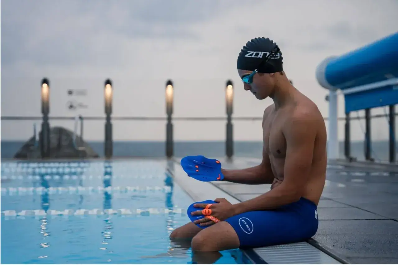 A swimmer sits by the pool, holding blue swim paddles, ready to learn how to use swim paddles for a stronger stroke.