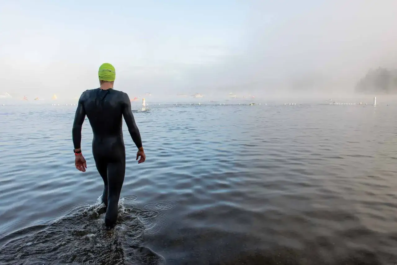 Athlete in wetsuit and cap enters misty water for a triathlon swim. Buoys mark the course.