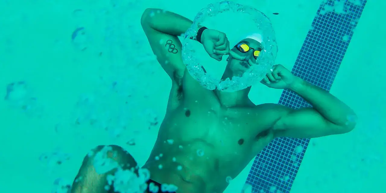 Swimmer exhales a bubble ring, demonstrating how to breathe in water. Olympic rings tattoo visible.