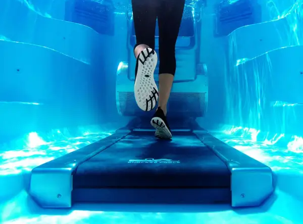 Underwater view of a person's legs and feet on a treadmill, performing aqua jogging in a bright blue pool.