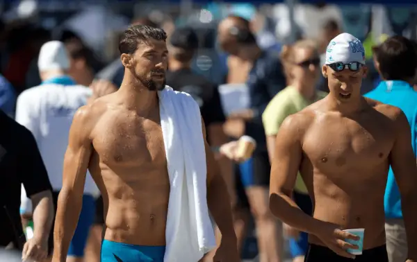 Two muscular men, one with a towel draped over his shoulder, showcase the lean physique of a swimmer body vs. a gym body.
