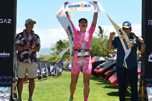 Triumphant athlete crosses the finish line at Hawaii 70.3, holding the banner high. Two men in leis congratulate her.