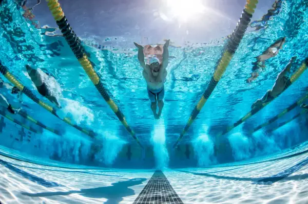 Underwater view of swimmers in a pool, with a focus on a swimmer performing butterfly stroke. The sun shines through the water, illuminating the lanes and the powerful stretches for swimmers.