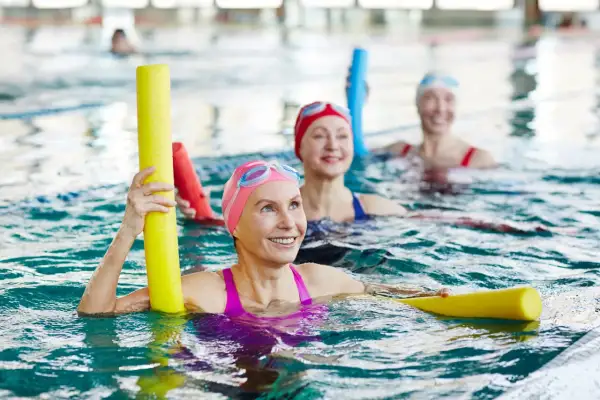 Three smiling fitness swimmers in a pool, holding yellow pool noodles.