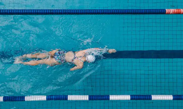 A lone swimmer glides through a pool lane, their back and arms creating splashes. The colorful swimsuit stands out against the blue water.