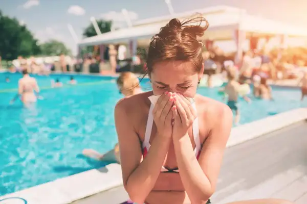 A woman with swimmer's sinusitis sneezes into a tissue by a crowded pool, unable to enjoy the summer day.