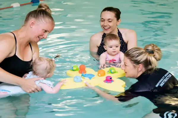 Moms and instructors in a pool with toddlers, playing with bath toys. The question of how many swim lessons does a toddler need is a common one for parents.