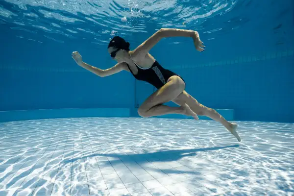 Underwater shot of a woman in a black swimsuit performing aqua aerobics. Her body is streamlined as she propels herself through the blue water.
