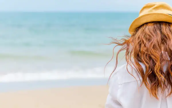 Woman with red hair in a straw hat on the beach, wondering how soon can I swim after coloring my hair.