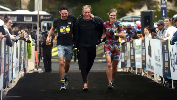 Three friends celebrate crossing the finish line of an Ironman relay. The woman in the colorful floral suit beams, while the two men, one in a black shirt with a race bib, share a laugh.