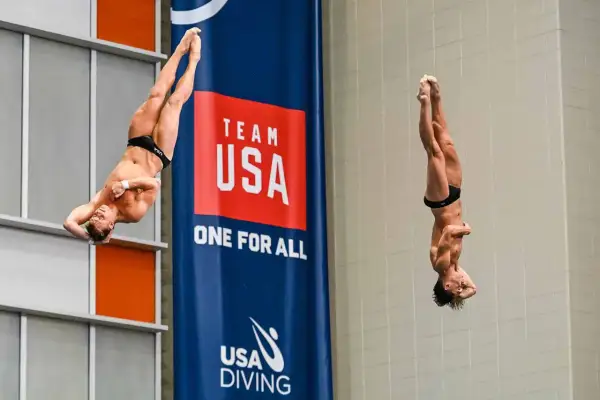 Two divers perform a synchronized high dive, suspended mid-air in a dramatic pose against a "Team USA" banner.