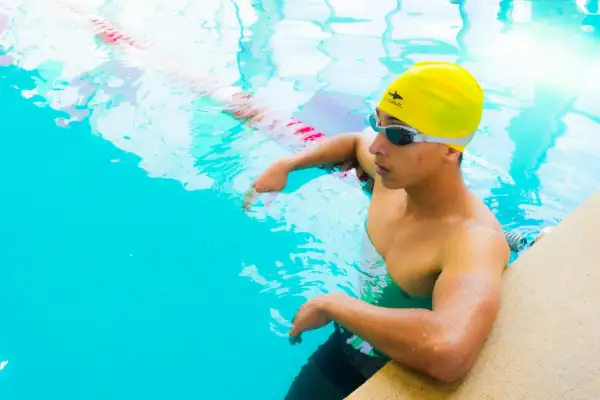 A young swimmer in a yellow cap and goggles rests by the pool edge, contemplating how much do swim instructors make.
