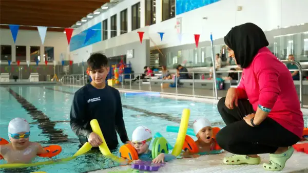 A swim instructor in a pink shirt and hijab coaches young children in a pool, demonstrating the rewarding nature of swim instructor jobs.