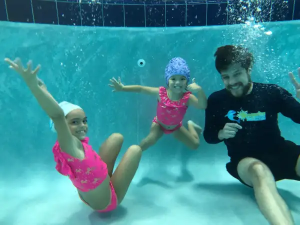 A swim instructor smiles while teaching two young girls how to teach toddler to hold breath underwater.