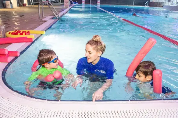 A smiling swim instructor in a blue shirt teaches two young boys in a pool. They are using pink pool noodles for support.