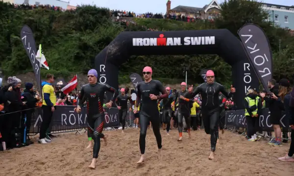Athletes in wetsuits run from the water under an "Ironman Swim" arch. The start of their race is a sandy beach, ready for the iron man swim challenge.