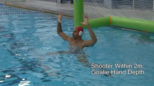 A water polo goalie in a red cap with hands raised, ready to block a shot.